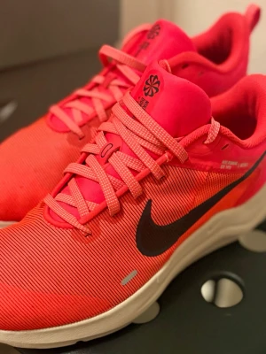 Nike rosa sneakers med svart swoosh - Snygga sneakers från Nike i en stark rosa färg med svart swoosh på sidan. Skorna har lågt skaft, klassisk snörning och ovandel i textil med diskreta ränder. Mellansulan är vit och yttersulan svart. Perfekta för dig som gillar sportig och färgstark stil.