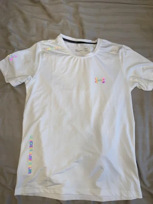 Vit t-shirt från Under Armour - Vit t-shirt från Under Armour med holografiska loggor och textdetaljer. T-shirten har rund halsringning och korta ärmar. Materialet känns sportigt och lätt, perfekt för en aktiv livsstil.