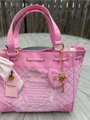 Juicy Couture daydreamer fairytale tote  - Superfin rosa handväska från Juicy Couture med quiltat mönster, guldfärgade detaljer och hjärtformat emblem med nyckelberlock. Väskan har dubbla handtag och dragskostängning. Materialet är en mix av mockaliknande tyg och konstläder.