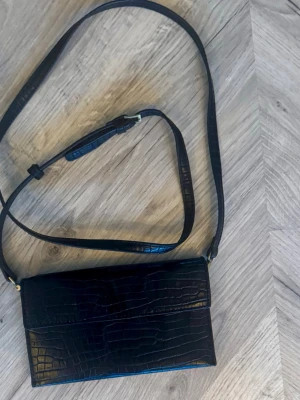 Svart axelväska i krokopräglat faux skinn från Åhléns  - Snygg svart axelväska med krokodilpräglat mönster och justerbar axelrem. Väskan har en stilren, rektangulär form och stängs med ett lock. Perfekt för dig som gillar trendiga accessoarer med lite edge. Innerfickan fastnade i dragkedjan så jag gjorde liten öppning för att komma åt fickan. Inget man ser utifrån. 