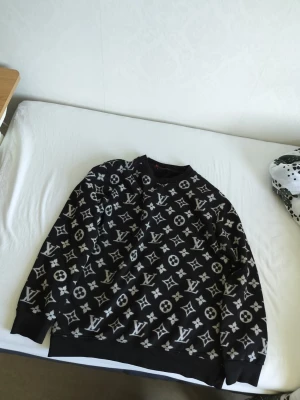 Louis Vuitton svart sweatshirt - Svart sweatshirt från Louis Vuitton med ikoniskt monogrammönster i vitt över hela tröjan. Rund halsringning och ribbade muddar vid ärmslut och nederkant. Perfekt för dig som gillar lyxiga streetwear-vibbar.
