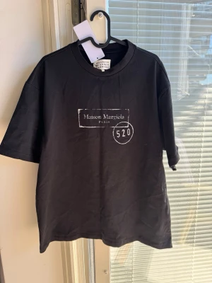 Maison Margiela svart t-shirt - Snygg svart t-shirt från Maison Margiela med vit logga och trycket '520' på bröstet. Klassisk rund halsringning och korta ärmar. Tillverkad i mjuk bomull för en skön känsla och avslappnad passform. Perfekt för dig som gillar stilrena designer.