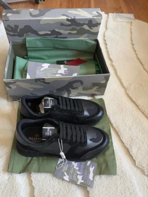 Svarta sneakers från Valentino Garavani - Snygga svarta sneakers från Valentino Garavani med grått camouflagemönster och svarta detaljer. Skorna har klassisk snörning, logga på plösen och är tillverkade i en mix av läder och textil. Kommer med originalkartong och dustbag.