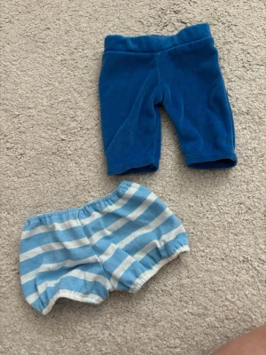 Blå och randiga shorts i bomull - Två par shorts, ett par helblå och ett par ljusblå/vit-randiga. Båda har resår i midjan och är gjorda i mjuk bomull, perfekta för dockor. De har en enkel och bekväm passform som passar till det mesta.