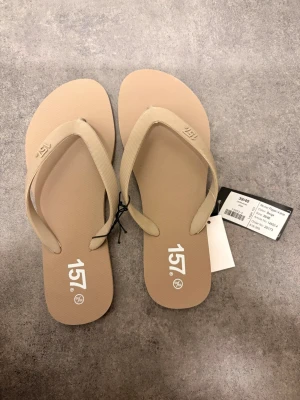 Beige flip-flops från 157 - Snygga beige flip-flops från 157 med klassisk design och breda remmar i matchande färg. Tillverkade i lätt och bekvämt syntetmaterial, perfekta för sommardagar. Diskret logga på fotbädden ger en clean look.