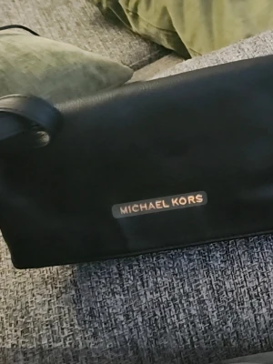 Svart handväska från Michael Kors - Snygg svart handväska från Michael Kors i slätt läder med guldfärgade detaljer och logotyp framtill. Väskan har ett praktiskt blixtlås och justerbar axelrem. Perfekt för dig som vill ha en stilren och klassisk accessoar.