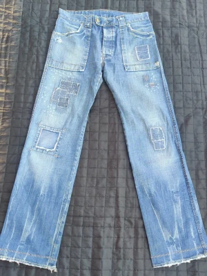 Blå patchwork design jeans med raka ben - Unika blå jeans med raka ben och patchwork-detaljer framtill och baktill. Jeansen har stora fickor fram och bak, slitna kanter och fransiga benslut. Tillverkade i denim med coola lagningar och slitningar för en trendig look. Viktigt att läsa: byxorna är i mycket gott skick MEN nertill slitna för dem var lite för långa för mig (se bilder) och baktill i stjärten är designen gjorda med två mindre hål! VIKTIGT LÄS DETTA!