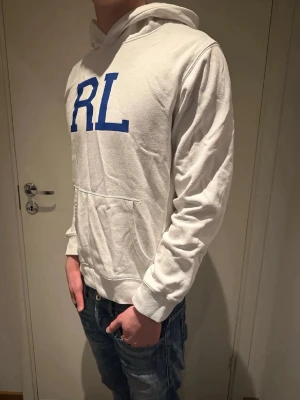 Vit hoodie från Ralph Lauren RL🤩🙌 - Vit hoodie med blå RL-tryck på bröstet från Ralph Lauren. Klassisk modell med huva och känguruficka framtill. Mjuk bomullskänsla och ribbade muddar vid ärmslut och midja. Perfekt för en avslappnad och stilren look. 