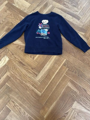 Mörkblå sweatshirt Polo Bear - Mörkblå sweatshirt från Ralph Lauren med Polo Bear-tryck framtill. Tröjan har rund halsringning, långa ärmar och ribbade muddar. Perfekt för en avslappnad och cool stil.