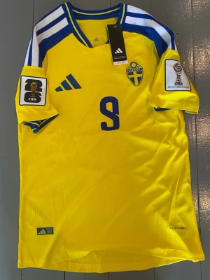 Sverige landslagströja Adidas gul - Gul Sverige landslagströja från Adidas med blå detaljer, nummer 9 på bröstet och officiella FIFA och VM märken på ärmarna. Tröjan har broderat landslagsemblem och är tillverkad i lätt funktionsmaterial (Climacool). Perfekt för fotbollsfans!