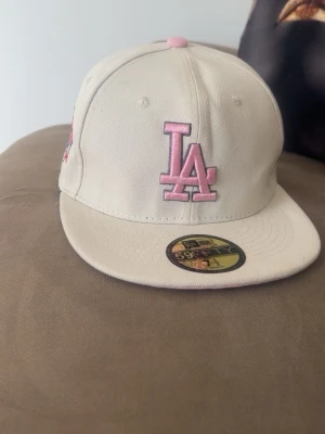 Beige LA New Era keps med rosa detaljer - Säljer en beige New Era 59Fifty keps med LA-logga i rosa framtill och broderade detaljer i rosa och grått på sidan. Kepsen har platt skärm och klassisk snapback-form. Perfekt för dig som vill ha en unik och snygg accessoar till din streetwear-look.