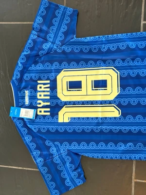 Blå Sverige fotbollströja Adidas - Säljer en blå Sverige fotbollströja från Adidas med gult tryck, nummer 18 och namnet Ayari på ryggen. Tröjan har gula ränder på axlarna, Sveriges landslagsemblem på bröstet och är tillverkad i lätt funktionsmaterial. Perfekt för fotbollsfans!