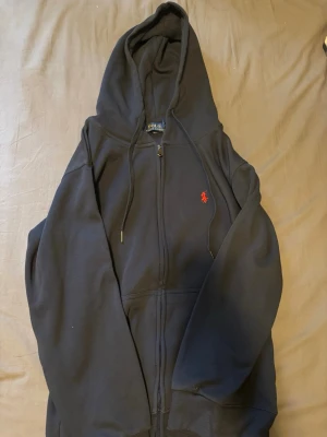 Marinblå hoodie från Polo Ralph Lauren - Snygg marinblå hoodie från Polo Ralph Lauren med dragkedja och huva. Klassisk röd broderad logga på bröstet och justerbara snören i huvan. 