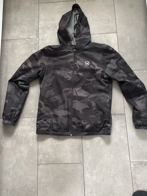 Svart camo windbreaker från Hollister - Cool svart vindjacka från Hollister med camo-mönster och huva. Jackan har dragkedja framtill, två sidofickor och en liten logotyp på bröstet. Insidan är fodrad med grått tyg för extra komfort. Perfekt för att hålla vinden borta under blåsiga dagar. Perfekt för dig som behöver en jacka till lite varmare väder. 