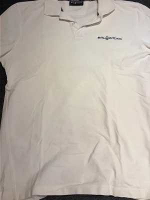 Vit pikétröja från Sail Racing - Klassisk vit pikétröja från Sail Racing med broderad logga på bröstet. Tröjan har krage, korta ärmar och knäppning framtill. Perfekt för en clean och sportig stil.