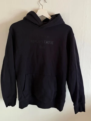 Svart hoodie Replay League 1981 - Svart hoodie från Replay med trycket 'PROPERTY OF REPLAY LEAGUE 1981' på bröstet. Klassisk modell med huva och stor magficka. Mjuk bomullskänsla och ribbade muddar vid ärmslut och nederkant. Perfekt för en avslappnad streetstyle-look.