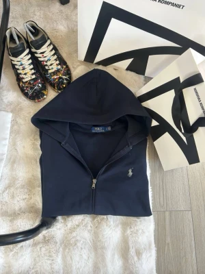 Mörkblå hoodie från Polo Ralph Lauren - Snygg mörkblå hoodie med dragkedja från Polo Ralph Lauren. Klassisk design med huva och den ikoniska loggan broderad på bröstet. Perfekt för en avslappnad och stilren look.