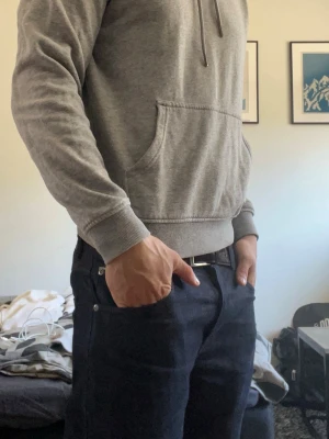 Grå basic hoodie med magficka - En stilren grå hoodie med huva och dragsnören. Klassisk magficka framtill och ribbade muddar vid ärmar och midja. Perfekt för en avslappnad look. Materialet känns mjukt och bekvämt, troligen bomullsblandning. Inga synliga loggor eller tryck.