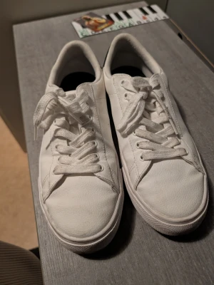 Vita låga sneakers med svart detalj - Stilrena vita sneakers med lågt skaft och klassisk snörning. Skorna har en svart detalj på hälen och är tillverkade i ett slätt material med gummisula. Perfekta för en clean och enkel look. Priset kan diskuteras och sänkas