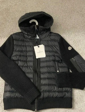 Svart hybridjacka från Moncler - Snygg svart hybridjacka från Moncler med vadderad front och huva samt stickade ärmar och sidor. Jackan har dragkedja, två fickor fram och Moncler-logga på ärmen. Perfekt för dig som vill ha en sportig och trendig look.