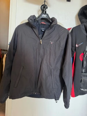 Svart vindjacka från Gant - Snygg svart vindjacka från Gant med dragkedja framtill och två fickor med dragkedja. Jackan har hög krage och diskret Gant-logga på bröstet. Perfekt för blåsiga dagar och enkel att matcha med olika outfits.