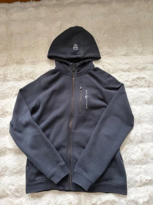 Svart hoodie från Peak Performance - Junior storlek 170. Snygg svart hoodie från sail racing med dragkedja hela vägen, huva och bröstficka med logga. Tillverkad i mjukt och bekvämt material, perfekt för en avslappnad stil. Klassisk passform med ribbade muddar och diskret logotyp på huvan.