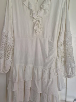 Vit volangklänning från Maje - Supersöt vit klänning från Maje med volanger, spetsdetaljer på ärmarna och v-ringning. Långärmad modell med flera lager volanger nertill.. Perfekt till sommaren eller studenten.