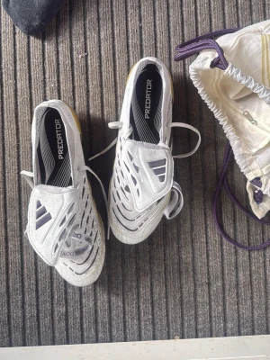 Vita Elite  Predator - Säljer ett par vita Adidas Predator fotbollsskor med svarta och lila detaljer. Skorna har snörning, högre plös och guld/guldfärgad sula med fasta dobbar. Tillverkade i syntetmaterial och designade för gräsplan. Perfekta för dig som vill ha kontroll och stil på planen.
