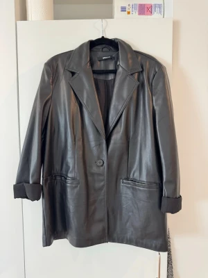 Oversized svart skinnjacka Gina tricot - Stilren oversized svart skinnjacka från Gina tricot med klassisk krage, en knapp framtill och två stora fickor. Jackan har uppvikta ärmslut och en rak, längre passform som ger en cool look.