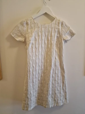 Vit kortärmad klänning med struktur - Snygg vit klänning med korta ärmar och rund halsringning. Klänningen har en strukturerad, quiltad yta med diskret rutmönster och är i en kort modell. Perfekt för dig som gillar stilrena och enkla plagg med en twist.  Inköpt i New York på en vintagebutik. Årets studentklänning?