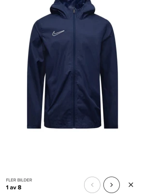 Mörkblå vindjacka från Nike - Snygg mörkblå vindjacka från Nike med huva och dragkedja framtill. Jackan har lång ärm, minimalistisk design och en vit Nike-logga på bröstet. Perfekt för blåsiga dagar och enkel att matcha med olika outfits.