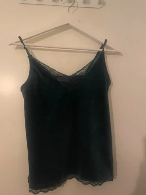 Mörkgrön sammetslinnetopp med spets - Mörkgrön pyjamaslinnetopp i mjuk sammet med tunna axelband och v-ringning. Spetsdetaljer längs halsringning och nederkant ger en extra touch. 