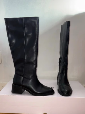 Svarta höga boots med klack - Stilrena svarta stövlar i äkta läder med hög skaft och låg bred klack. Klassisk design med rundad tå och diskreta sömmar längs skaftet. Perfekta för att lyfta vilken outfit som helst. Använda en gång. Köpta från NAKD.