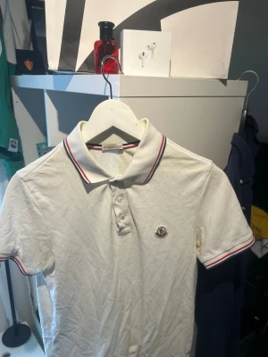 Vit Moncler pikétröja med rödblå detaljer - Klassisk vit pikétröja från Moncler med korta ärmar och krage. Tröjan har röda och blå ränder på kragen och ärmsluten samt Moncler-logga på bröstet. Perfekt till en clean och sportig stil. Den är i XC men skulle passat bra på någon som har S också