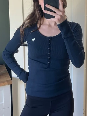Mörkblå ribbad topp Abercrombie - Mörkblå ribbad topp från Abercrombie & Fitch med lång ärm och rund halsringning. Dekorativa knappar framtill och liten vit logga på bröstet. Sitter tight och är perfekt för en enkel men snygg look.