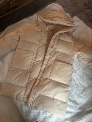 Beige pufferjacka med huva - Säljer en beige pufferjacka med huva och stora, rejäla fickor framtill. Jackan har dragkedja och tryckknappar, quiltad design och är perfekt för kalla dagar. Oversized look och riktigt mysig att bära under vintern. Har en fläck på luvan som inte syns när man har på sig den.