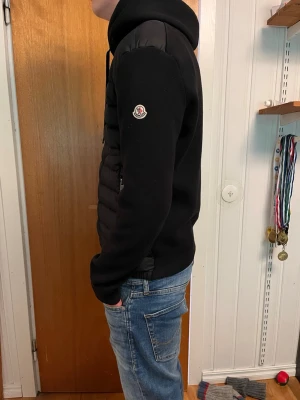 Svart cardigan från Moncler - Säljer en svart pufferjacka från Moncler med huva och dragkedja framtill. Jackan har quiltad front och släta ärmar, samt Moncler-logga på vänster ärm. Perfekt för kalla dagar och riktigt snygg streetstil.