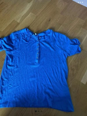Blå kortärmad topp med knappar - En blå topp med korta ärmar och knappar framtill. Toppen har en enkel och avslappnad passform, perfekt för en chill look. Materialet känns mjukt och lätt, och knapparna ger en snygg detalj till framsidan.