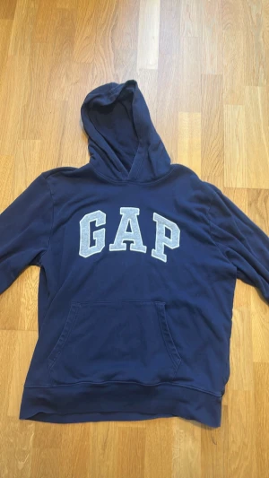 Mörkblå hoodie från GAP - Mörkblå hoodie från GAP med stor logga i vitt och grått framtill. Klassisk känguruficka och huva. Mjuk bomullsblandning som är skön att ha på sig. Perfekt för en avslappnad och stilren look.