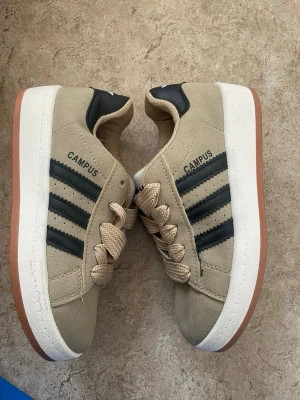 Beige Adidas Campus sneakers - Snygga beige Adidas Campus sneakers med klassiska svarta ränder på sidan och vit sula. Ovandelen är i mockaliknande material och skorna har beigea snören samt gummisula i brunt. Perfekta för en avslappnad och trendig stil.
