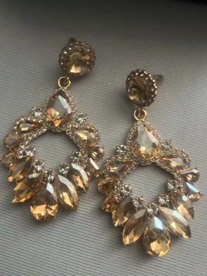 Guldiga statement-örhängen med kristaller - Säljer ett par stora, guldiga statement-örhängen med gnistrande, facetterade kristaller i droppform och runda stenar. Örhängena har en lyxig känsla med många detaljer och glittrande inslag. Perfekta för att sticka ut och ge en glamorös touch till din look.