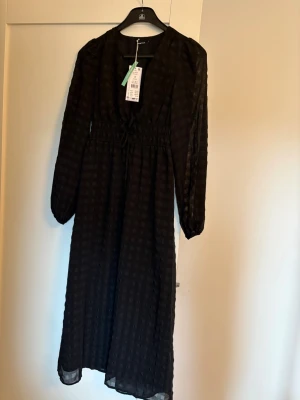 Svart långklänning från Gina Tricot - Svart djupt v-ringad långklänning från Gina. Klänningen har långärmade ärmar, knyt under bysten och är tillverkad i ett lätt och luftigt material som ger en snygg siluett. Har innerkjol. Aldrig använd. Nypris 599 kr.