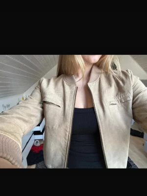Beige bomberjacka i mockaimitation - Snygg beige bomberjacka i mockaimitation med ribbade muddar och krage. Jackan har två bröstfickor med dragkedja och klassisk bomberpassform. Perfekt för dig som vill ha en stilren och trendig look.