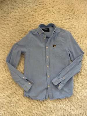 Ljusblå skjorta från Lyle & Scott - Klassisk ljusblå skjorta från Lyle & Scott med button-down krage och bröstficka med den ikoniska gula örnloggan. Skjortan har vita knappar och är tillverkad i bomull, perfekt för en stilren och avslappnad look.