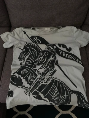 Vit Burberry t-shirt med tryck - Snygg vit t-shirt från Burberry med stort svart tryck av riddare och flagga med texten 'Prorsum' på framsidan. Klassisk rund hals och korta ärmar. Tillverkad i mjuk bomull för en skön känsla.