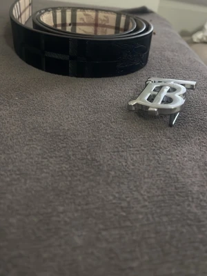 Burberry rutigt bälte med silverdetalj - Säljer ett klassiskt Burberry bälte i skinn med ikoniskt rutigt mönster i beige, svart och vinrött. Bältet har en stor silverfärgad BB-logga som spänne. Perfekt accessoar för att lyfta din outfit.