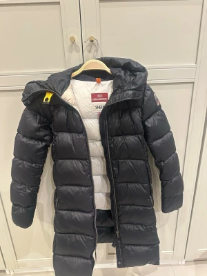 Svart lång dunjacka Parajumpers - Snygg svart lång dunjacka från Parajumpers med huva och dragkedja framtill. Jackan har vita detaljer på insidan och klassisk gul Parajumpers-detalj vid halsen. Behövs kemtvätt därav de priset. 