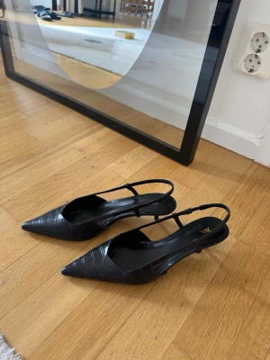 Svarta pumps med krokodilmönster från  Zara - Snygga svarta pumps från Zara med spetsig tå och slingback-rem. De har ett krokodilmönster i konstläder och låg klack. Knappt använda och fint skick❤️