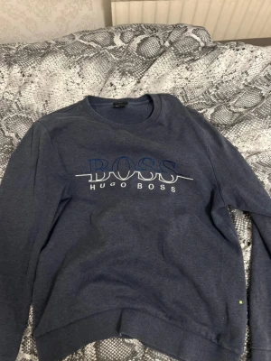 Blå sweatshirt från Hugo Boss - Säljer en blå sweatshirt från Hugo Boss med stor BOSS-logga broderad på bröstet. Tröjan har rund halsringning, långa ärmar och ribbade muddar vid ärmslut och nederkant. Perfekt för en avslappnad och stilren look.