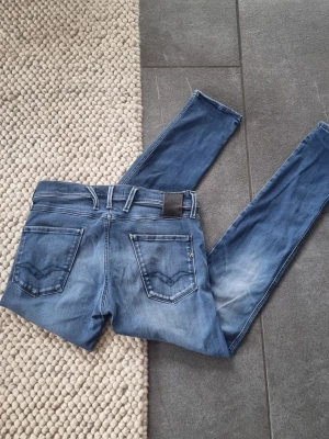 Replay Anbass - Snygga blå slim fit jeans från Replay med klassisk femficksdesign och diskreta slitningar framtill. Jeansen har raka ben, normal midja och detaljerade bakfickor med sömmar. Perfekta för en avslappnad och trendig look.